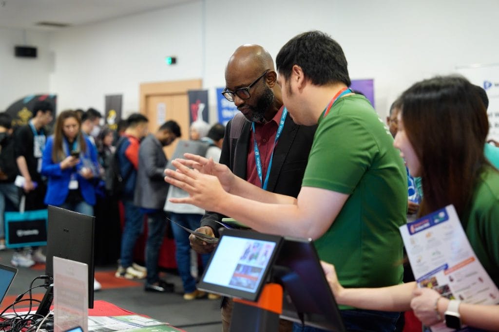 People interact at a tech event; one person gestures while others observe a display with information materials around.