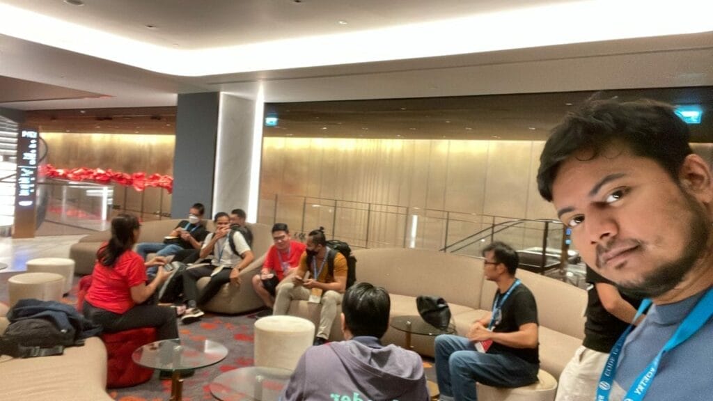A group of people sit and talk on curved couches in a modern lounge area; one person in the foreground faces the camera, taking a selfie.