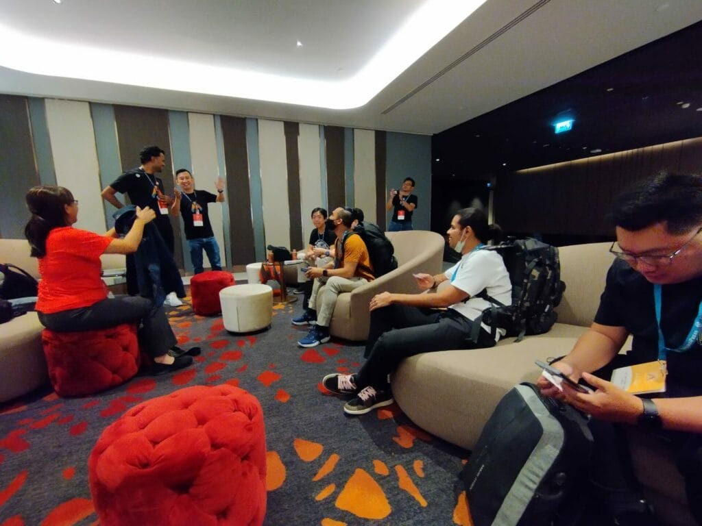 A group of people with conference badges sit and stand in a modern lounge area, some talking and gesturing, others using their phones or laptops.