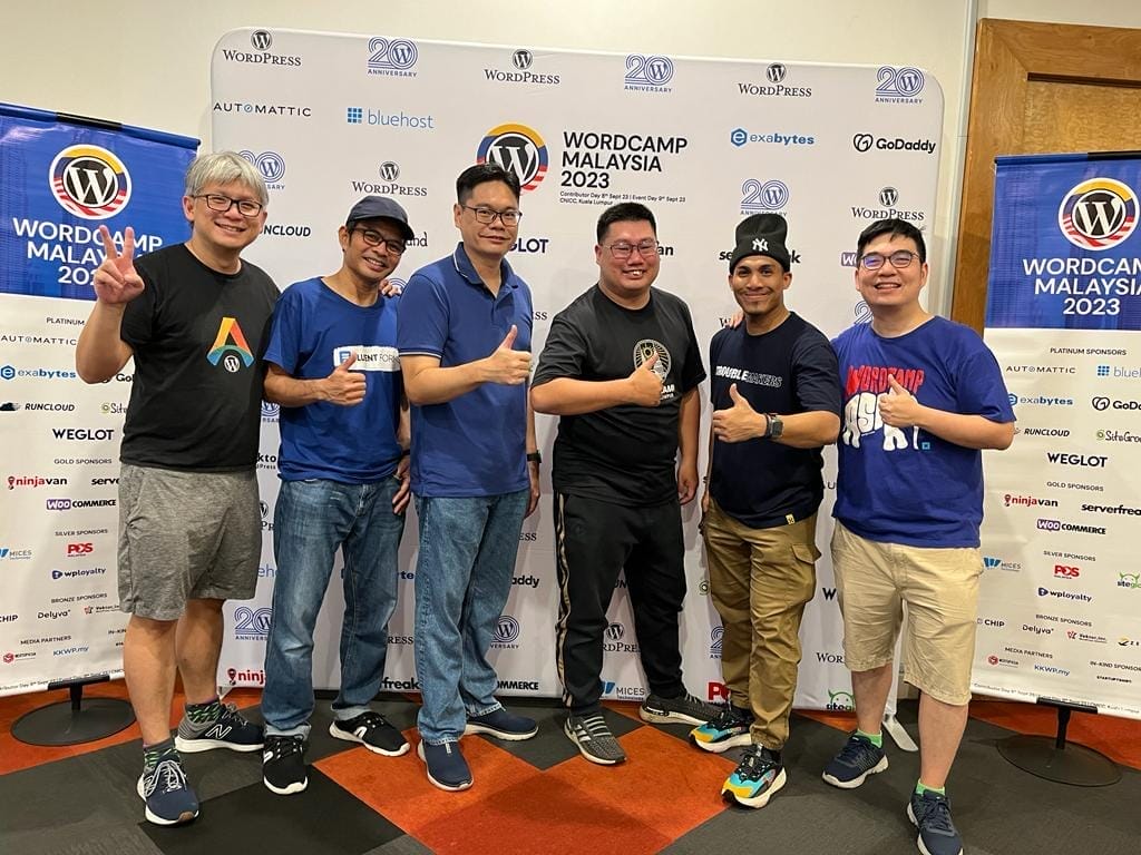 Six men standing side by side, smiling and giving thumbs up at WordCamp Malaysia 2023, with a branded backdrop displaying sponsor logos behind them.