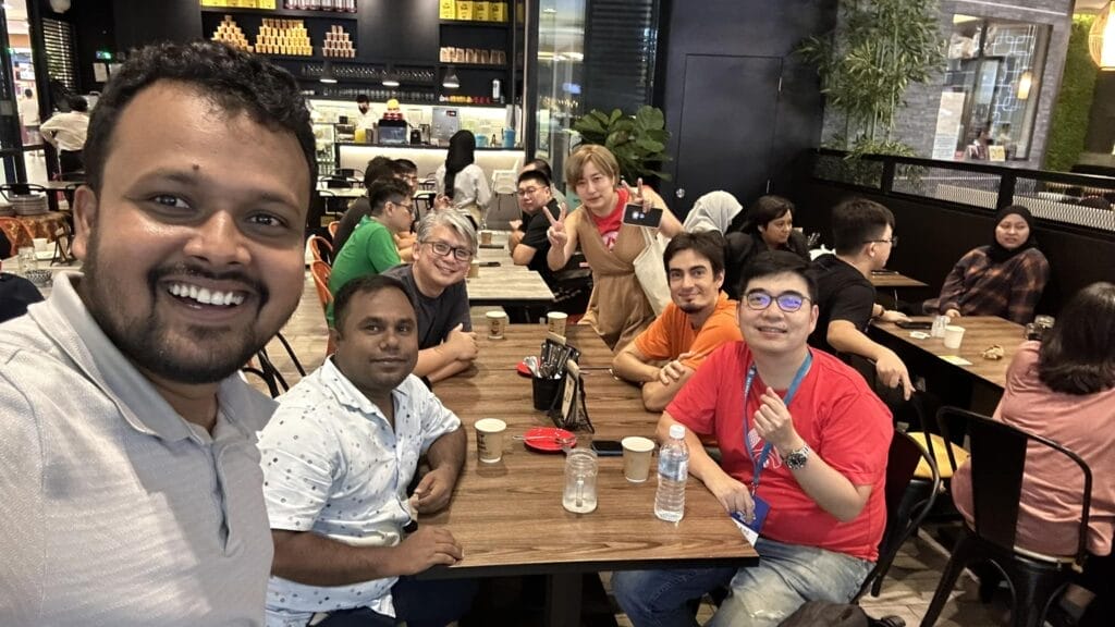 A group of six people sit and smile for a selfie at a café, with drinks and cups on the table; other patrons are visible in the background.