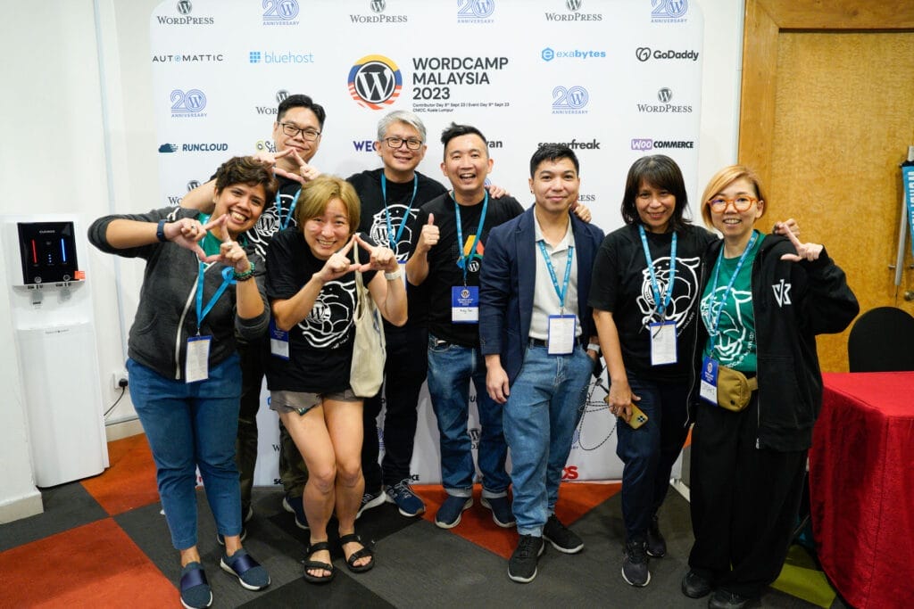 A group of eight people pose and smile in front of a WordCamp Malaysia 2023 backdrop, wearing event badges and branded t-shirts.