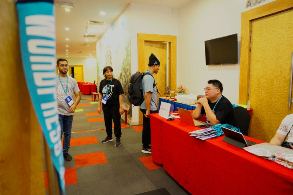 Several people stand near a registration desk with red tablecloths at an indoor event. A staff member sits behind the desk with a laptop and registration materials.