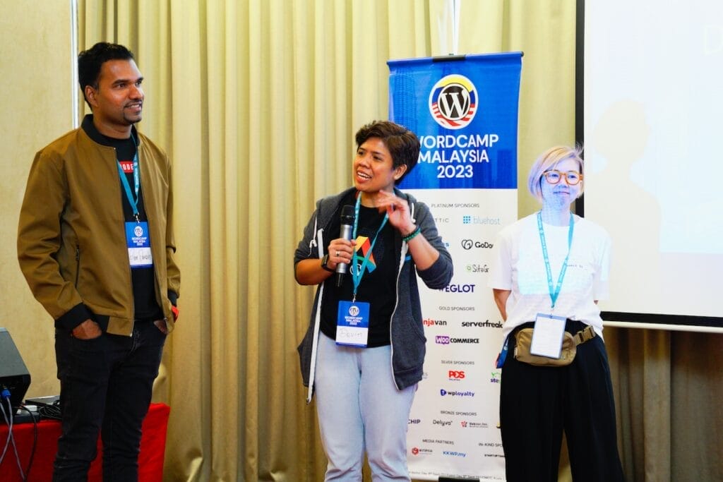 Three people stand in front of a WordCamp Malaysia 2023 banner; one person is speaking while holding a microphone, and the other two are listening.