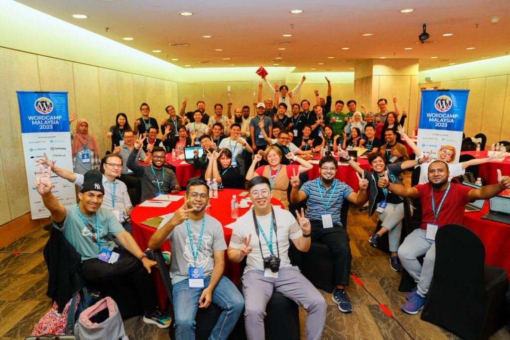 A large group of people sit and stand around tables at a WordCamp Malaysia 2023 event, smiling and making peace signs for a group photo indoors.
