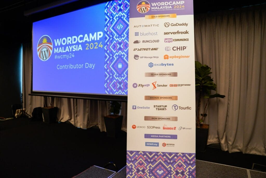 A standing banner displays sponsors for WordCamp Malaysia 2024 Contributor Day, with an event screen in the background and some plants beside the stage.