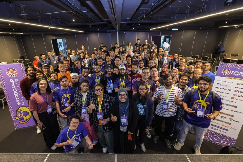 A large group of people pose for a photo on stage at WordCamp Malaysia 2023, with event banners and sponsor logos visible on both sides.