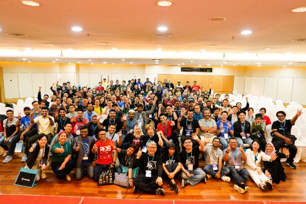 A large group of people pose for a group photo in a conference hall, many smiling and waving at the camera on a wooden floor with rows of chairs and bright overhead lights.