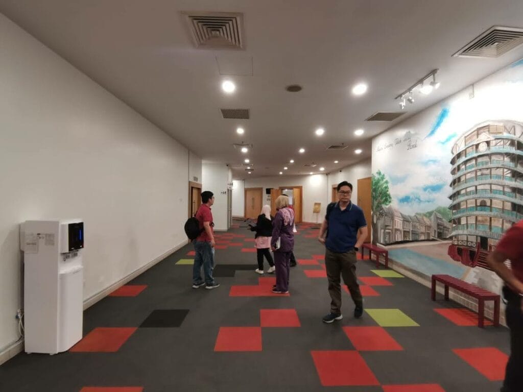 Several people stand and walk in a hallway with red and black checkered flooring, white walls, benches, a water dispenser, and a large mural painting on the right wall.