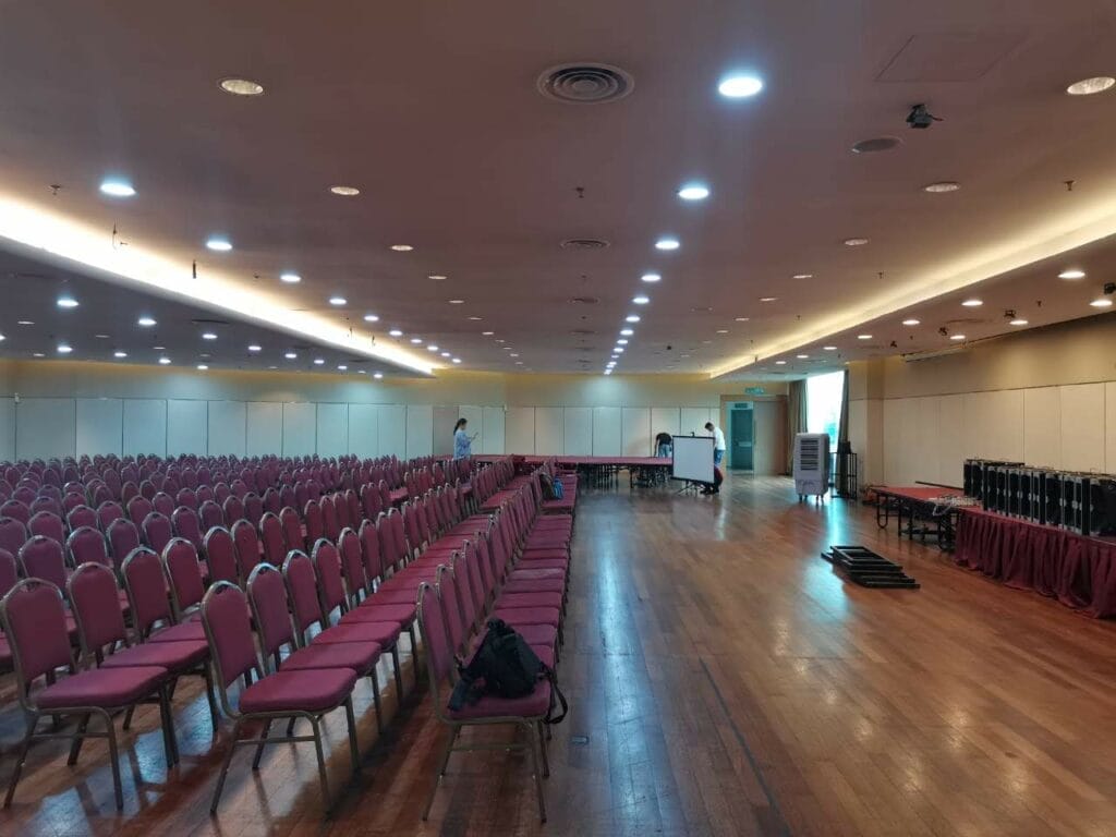 A large empty conference room with rows of red chairs, a wooden floor, and a few people setting up tables at the far end.