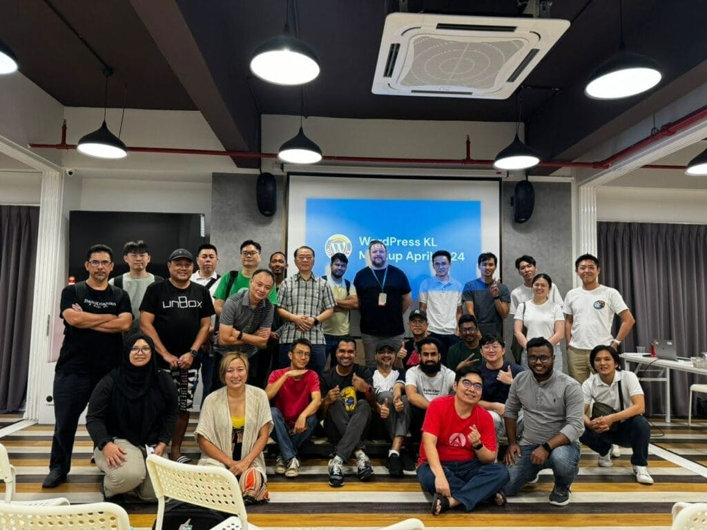 A group of people posing and smiling for a photo in front of a screen that reads "WordPress KL Meetup April 2024" in a modern indoor setting.
