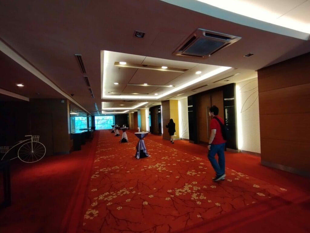 A hallway with red carpet, ceiling lights, cocktail tables covered with white cloth, and two people walking; a decorative bicycle is visible on the left.