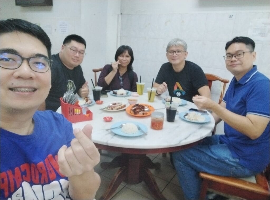 Five people sit around a round table with plates of food and drinks, smiling at the camera in a casual dining setting.