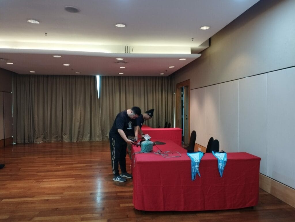 Two people arrange items on a red-covered table in a room with wooden floors, chairs, and tan curtains. Blue lanyards are laid out on the table.