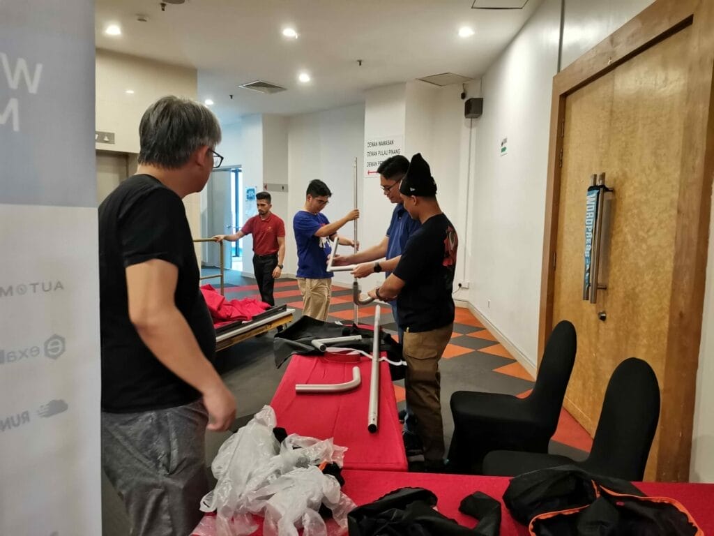 Four people assemble equipment and handle materials on tables and chairs in an indoor room with white walls and a patterned floor.