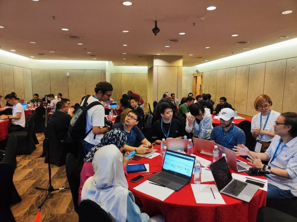 People are seated at round tables with laptops and notepads, engaged in discussion in a conference room setting.