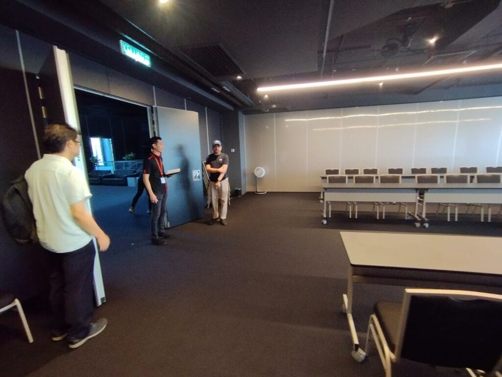 Four people stand and converse in a modern, dimly lit meeting room with empty chairs and tables arranged on the right side.
