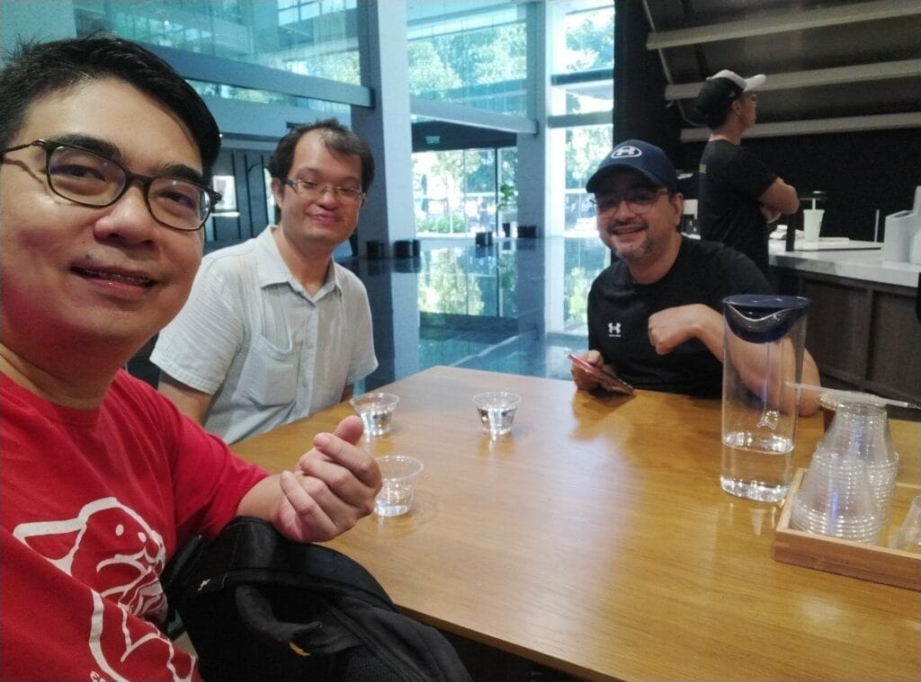 Three men sit at a wooden table with water glasses and a pitcher inside a modern building; one man takes a selfie.