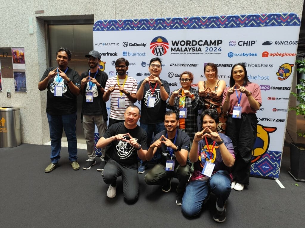 Ten people pose in front of a WordCamp Malaysia 2024 banner, making hand signs and wearing event badges.