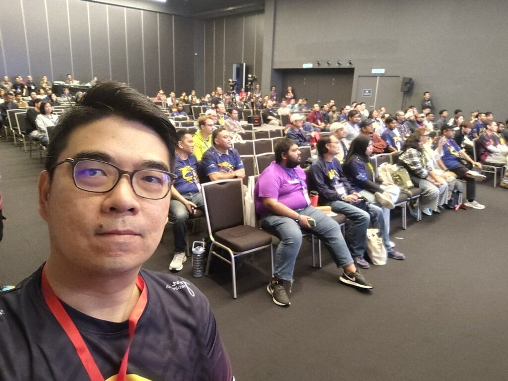 A man takes a selfie in front of an audience seated in rows at an indoor event or conference.