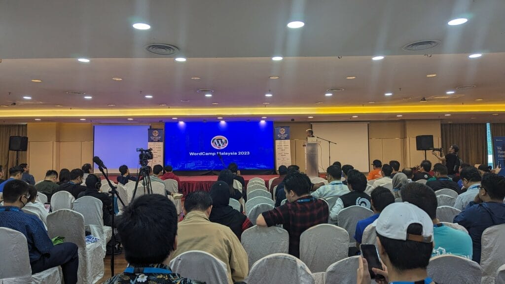 A speaker presents on stage at WordCamp Malaysia 2023 to an audience seated in a conference room.
