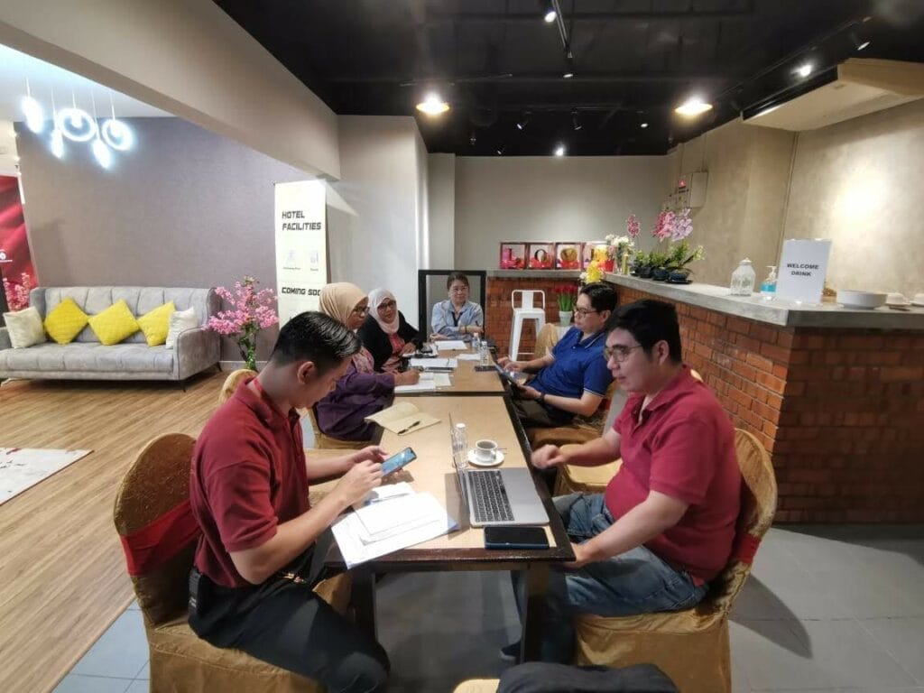 Six people sit around a table in a meeting room with laptops, papers, and phones; the space has modern decor with brick and floral elements.