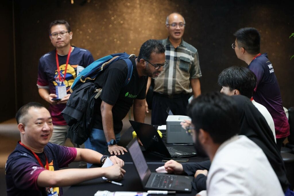 A group of people interact around a table with laptops during an indoor event; some are seated while others stand and converse.