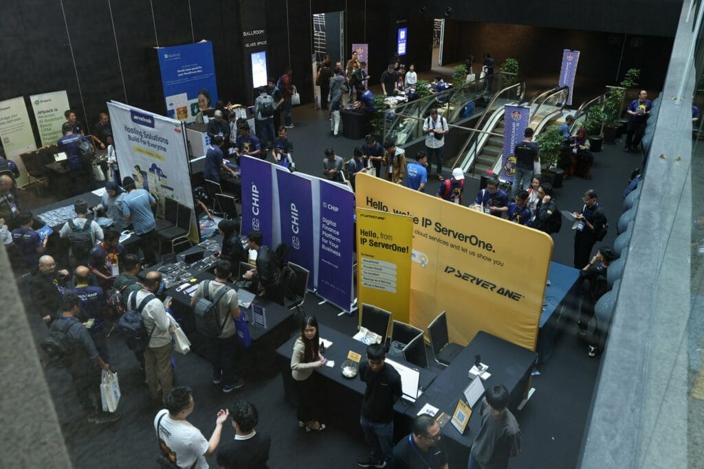 A busy tech conference hall with attendees interacting at exhibition booths, networking, and walking near escalators; banners and computers are visible.