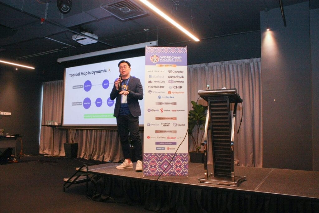 A man gives a presentation on "Topical Maps in Dynamic" at a conference, standing next to a podium with a banner displaying sponsor logos.