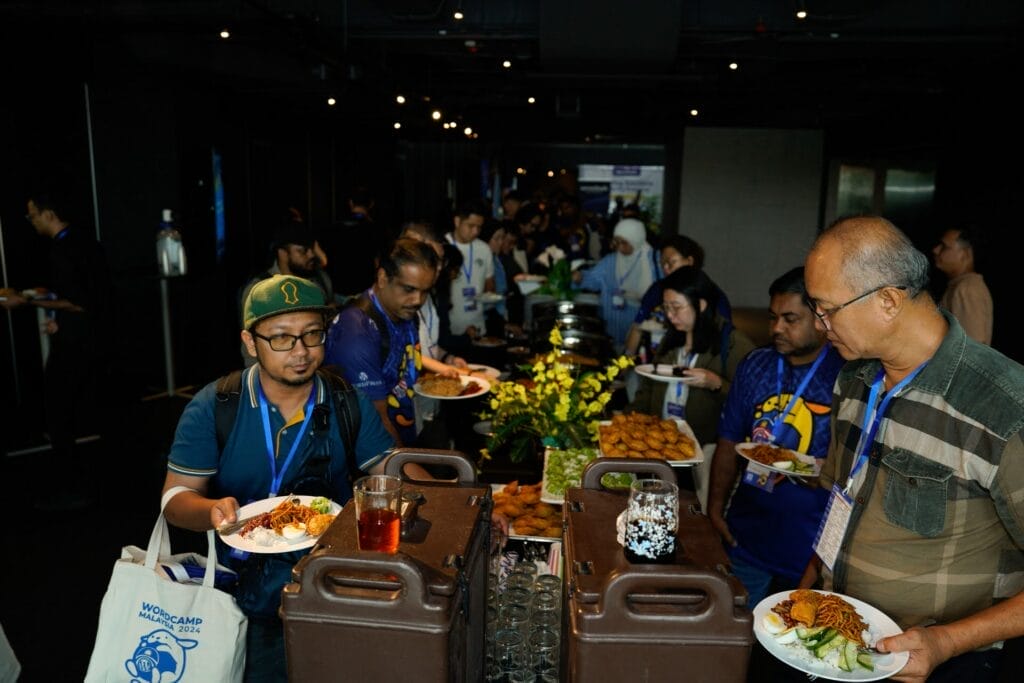 People serve themselves food and drinks from a buffet table at an indoor event, with others in the background holding plates and wearing attendee lanyards.