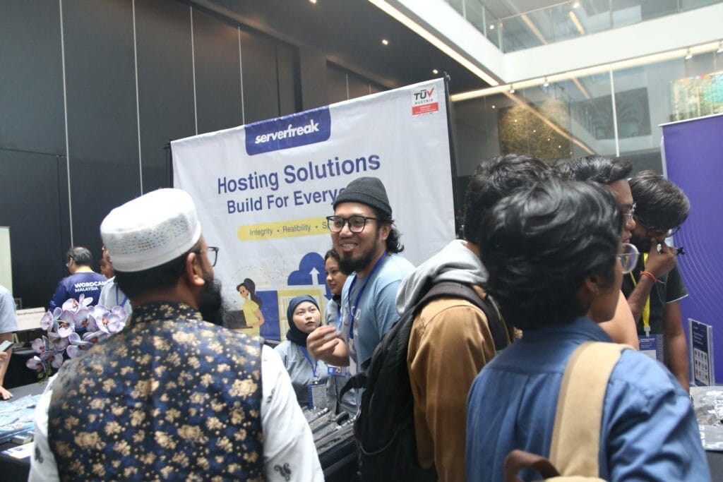 A group of people stand and talk at a tech expo booth with a "Hosting Solutions Build For Everyone" banner in the background.