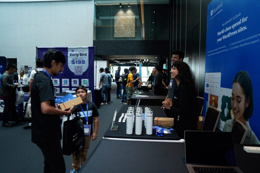 People interact at a tech expo booth with promotional materials and banners, while others walk around the event space in the background.