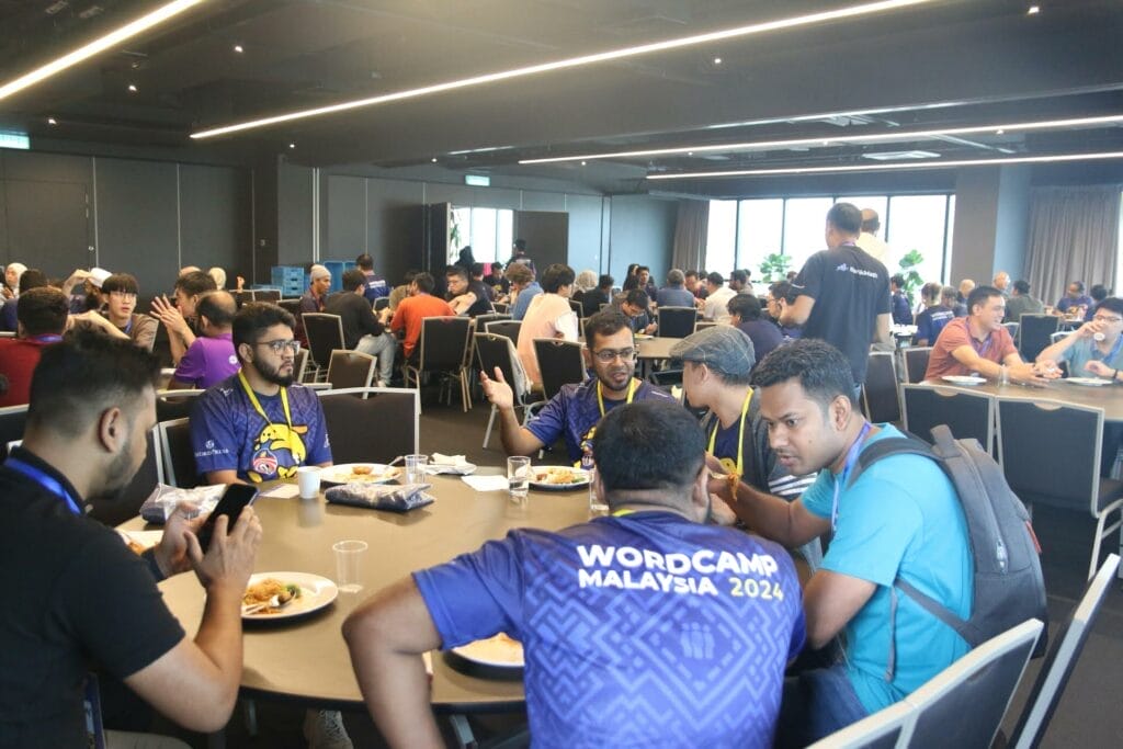 A large group of people sit at round tables eating and conversing at an indoor event; some shirts read "WordCamp Malaysia 2024.