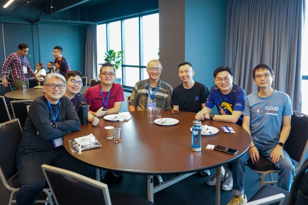 Seven men sit around a round table with food and drinks at a professional event in a modern indoor setting, all smiling at the camera.