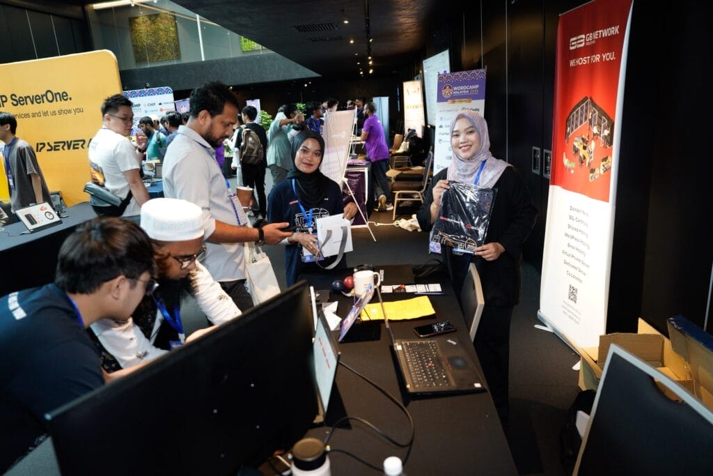 A group of people interact at a booth during a tech event; one woman in front smiles and holds a prize, while others are gathered around a table with laptops.