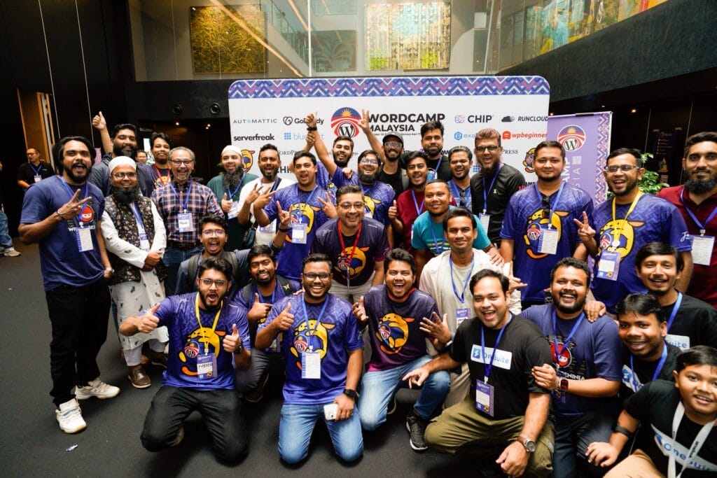A group of people pose and smile for a photo in front of a WordCamp Malaysia event banner indoors.