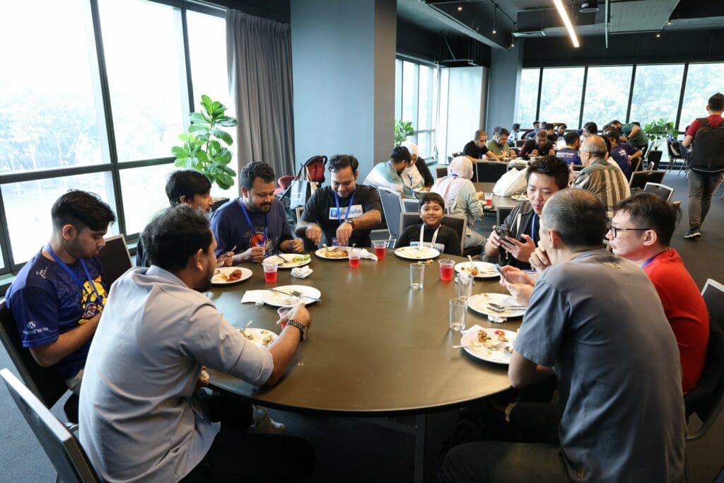 A group of people sit around a large round table eating food, with others dining at nearby tables in a brightly lit room with large windows.