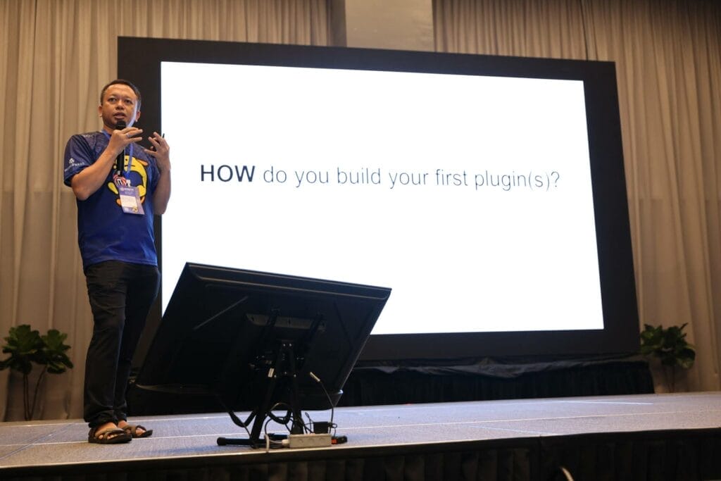 A speaker stands on stage next to a large screen displaying the text "HOW do you build your first plugin(s)?" at a presentation.