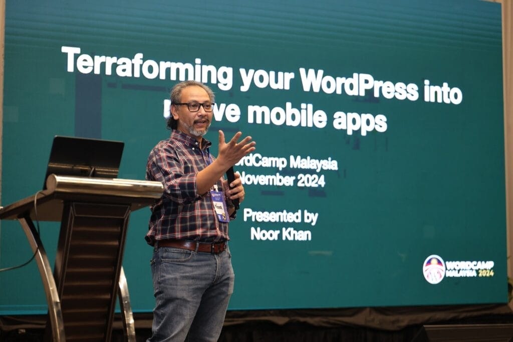 A man speaks on stage in front of a large screen displaying a presentation titled "Terraforming your WordPress into native mobile apps" at WordCamp Malaysia 2024.