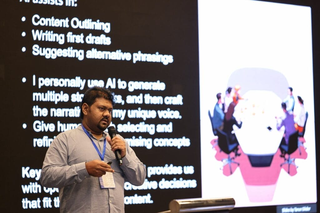 A man with a lanyard speaks in front of a screen displaying text and an illustration of people sitting around a conference table.