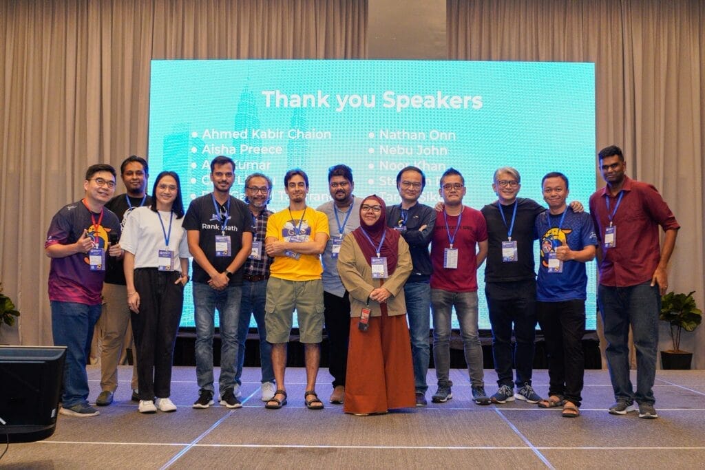 A group of thirteen people stands on a stage in front of a screen displaying a "Thank you Speakers" message and a list of names.