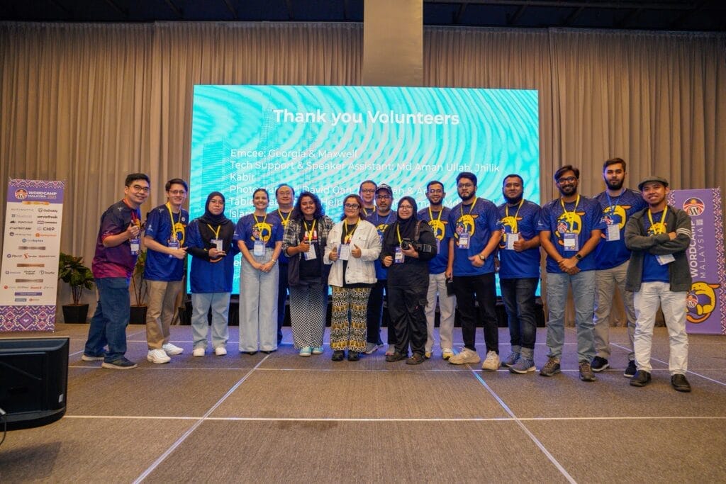 A group of people stand on a stage with plaques and medals, posing for a photo in front of a screen that reads "Thank you Volunteers.