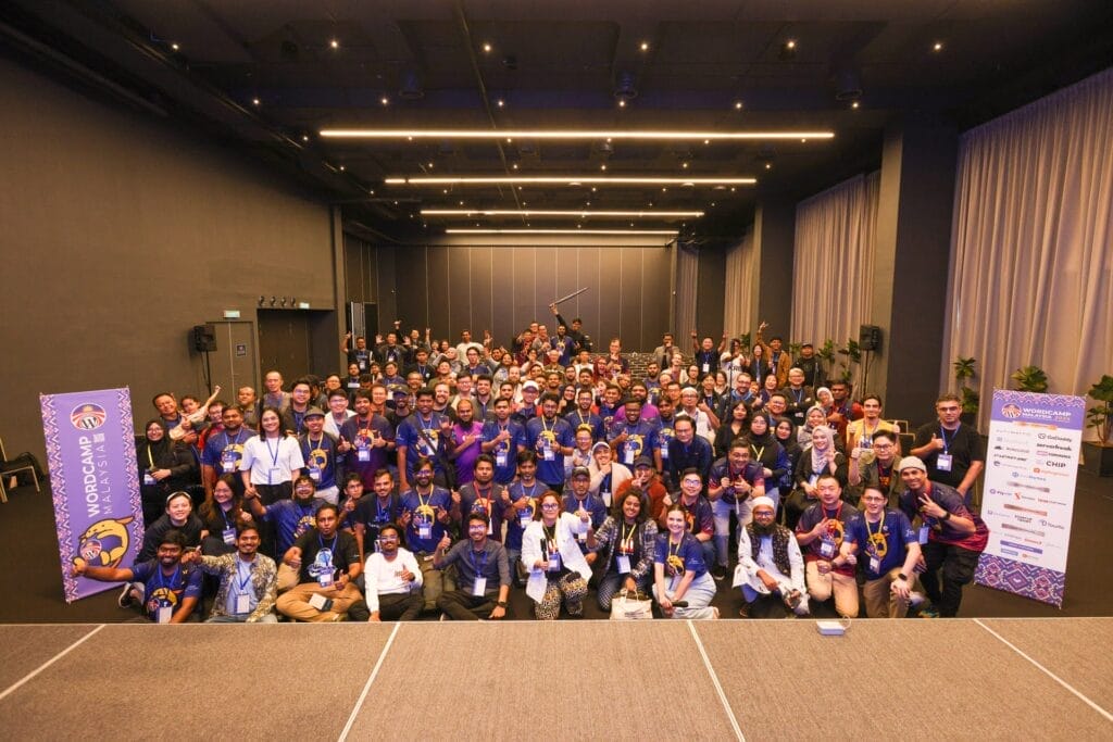A large group of people pose together for a photo at a conference, standing and sitting in rows inside a spacious event hall.