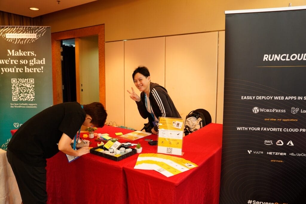 Two people at a booth with promotional materials; one is writing, and the other stands behind the table smiling and making a peace sign. Banners advertise tech services in the background.