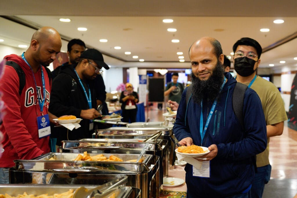 A group of people serve themselves food from a buffet line at an indoor event; one man at the front holds a plate and looks at the camera.