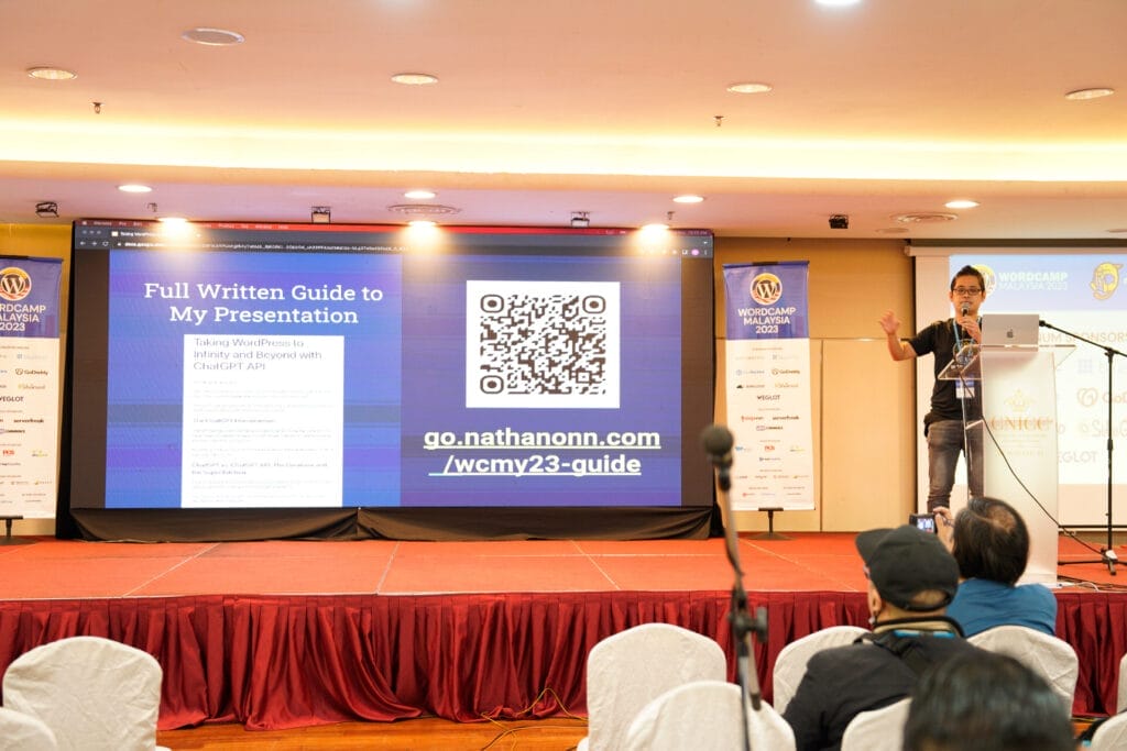 A speaker presents at a conference, pointing to a large screen displaying a QR code, a web link, and presentation slides on the stage. Audience members are seated in front.