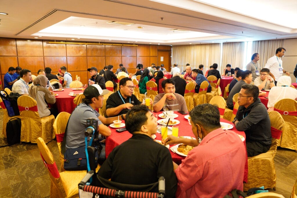 A large group of people sits at round tables in a banquet hall, eating and conversing during a meal event.
