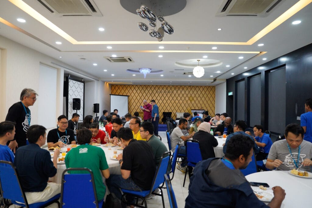 A group of people sit at round tables eating and conversing in a brightly lit conference room with modern decor.