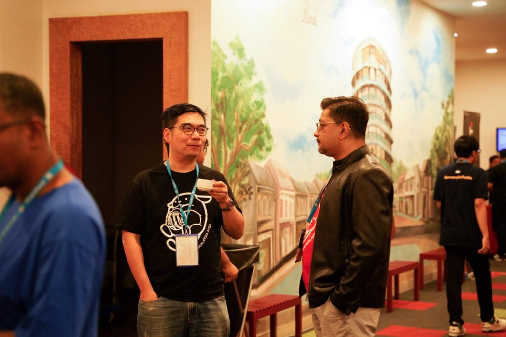 Two men stand and converse in front of a mural featuring the Leaning Tower of Pisa at an indoor event. Other people are seen in the background.