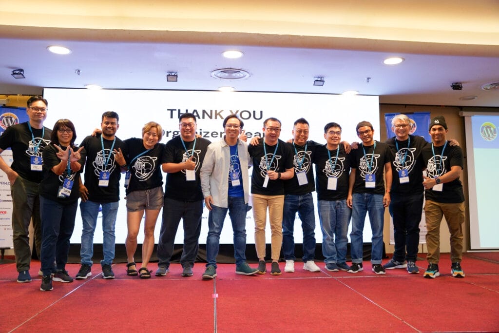 A group of twelve people stand on a stage in matching black event t-shirts, smiling and posing for a photo in front of a screen that reads "THANK YOU Organizer Team.